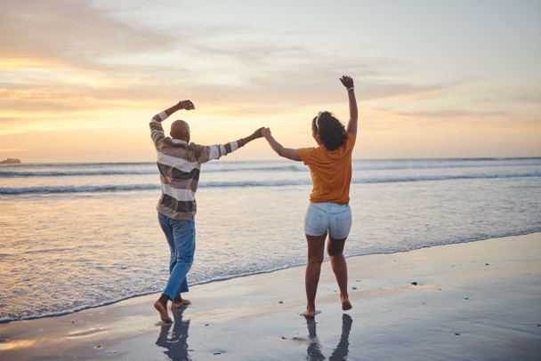 Love, travel and happy couple at beach enjoying summer vacation or fun honeymoon at sunset while holding hands and being playful. Laughing, energy and seaside holiday with black man and woman at sea. - Photo, Image
