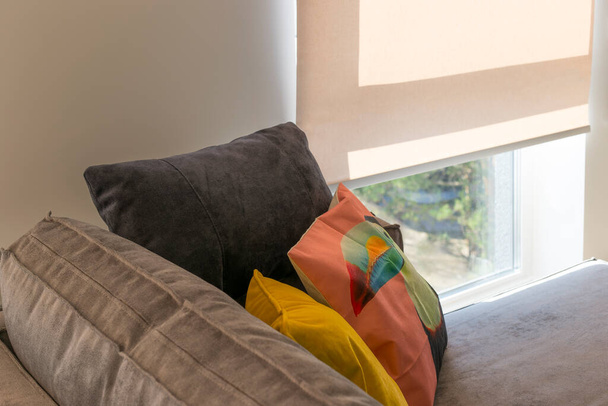 A sofa with colorful pillows in the room near a window with roller blinds. Roller shades on large window to the floor in the interior. Sunny day. Selective fokus. - Photo, Image