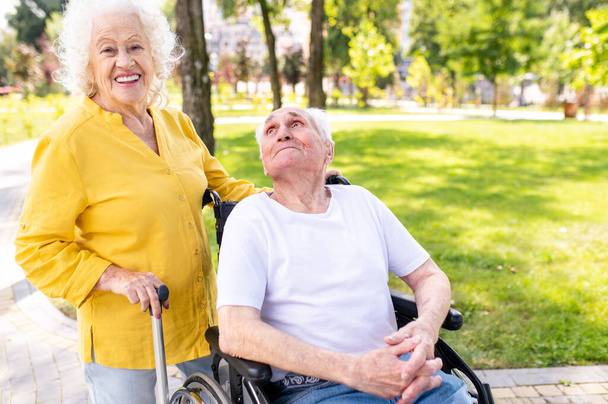 Beautiful senior couple with disability outdoors - Old people in the age of 60, 70, 80 having fun and spending time together, concepts about elderly, seniority, healthcare and wellness aging - Фото, изображение