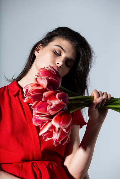 Atractiva morena joven en vestido rojo sosteniendo cerca de ramo de tulipanes rojos, fondo gris aislado. - Foto, imagen