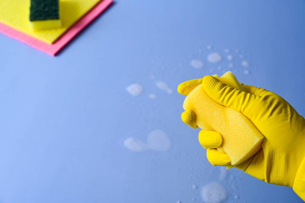 Mano con guante de goma amarillo en sostiene una esponja de limpieza sobre un fondo azul. - Foto, imagen