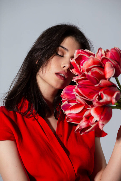 Приваблива брюнетка молода жінка з макіяжем в червоній сукні пахне букетом червоних тюльпанів, на сірому фоні
. - Фото, зображення