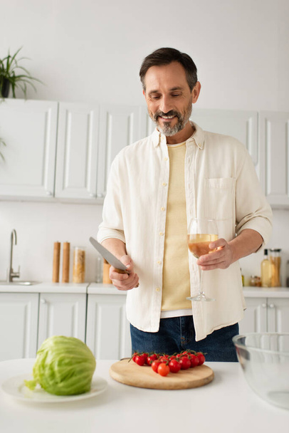 happy bearded man holding knife and wine glass near fresh lettuce and ripe cherry tomatoes - Fotoğraf, Görsel