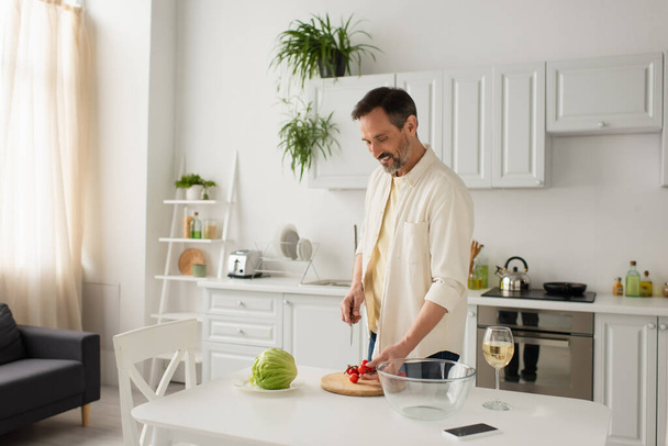 smiling man holding knife near cherry tomatoes and fresh lettuce in kitchen - Φωτογραφία, εικόνα