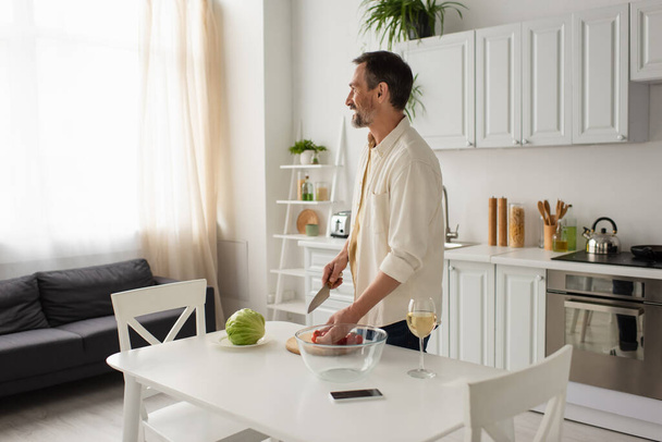 hombre barbudo feliz mirando hacia otro lado mientras está de pie cerca de verduras frescas y teléfono inteligente con pantalla en blanco en la cocina - Foto, Imagen