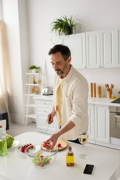 человек в белой рубашке готовит свежий овощной салат возле бутылок с маслом и бокалом вина - Фото, изображение