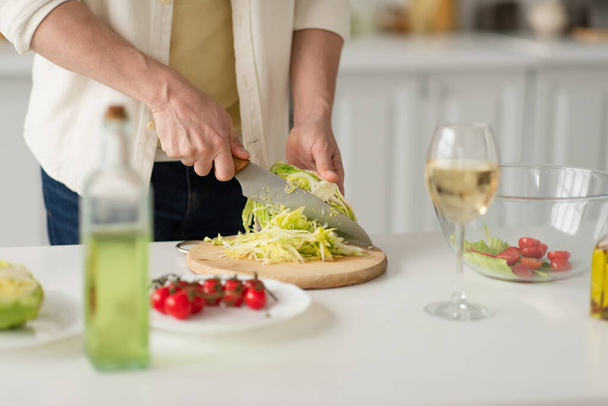 cropped view of man cutting lettuce near cherry tomatoes and blurred glass of white wine - Photo, image