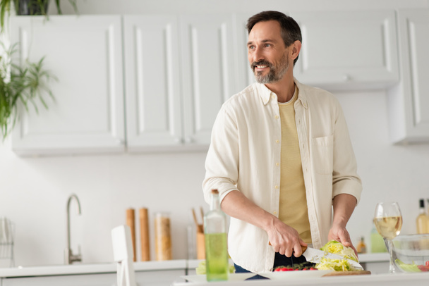 bearded man smiling and looking away while cutting lettuce near glass of white wine - Photo, Image