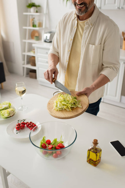 osittainen näkymä hymyilevästä miehestä valmistamassa salaattia salaatilla ja kirsikkatomaateilla älypuhelimen ja viinilasin lähellä - Valokuva, kuva