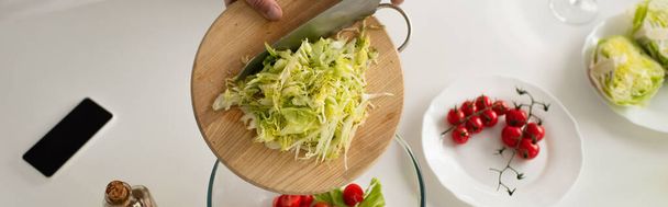 top view of cropped man holding chopping board with cut lettuce near cherry tomatoes and mobile phone with blank screen, banner - Photo, Image