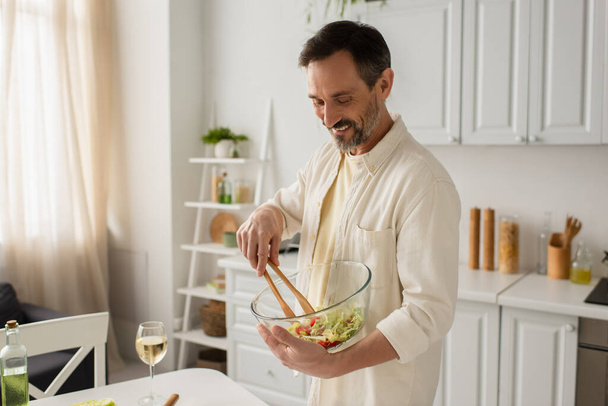 веселий чоловік, що змішує овочевий салат з дерев'яними щипцями біля келиха білого вина на кухні
 - Фото, зображення