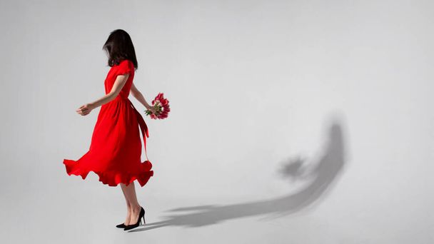 Πλήρης επιμήκυνση της εικόνας μιας μελαχρινής νεαρής γυναίκας με κόκκινο φόρεμα που κρατά ένα μπουκέτο τουλίπες και περιστροφές, απομονωμένο γκρι φόντο. Χώρος για κείμενο. - Φωτογραφία, εικόνα