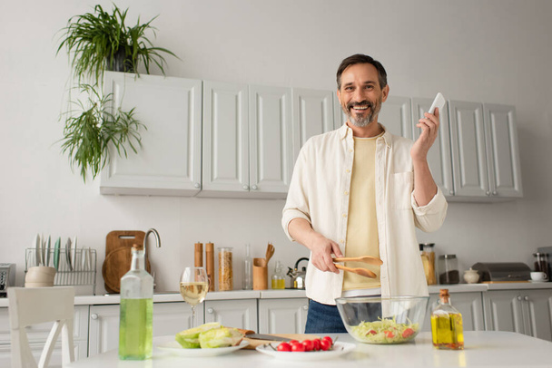 happy man with smartphone and wooden tongs looking at camera near fresh vegetables and bottles with oil - Fotoğraf, Görsel