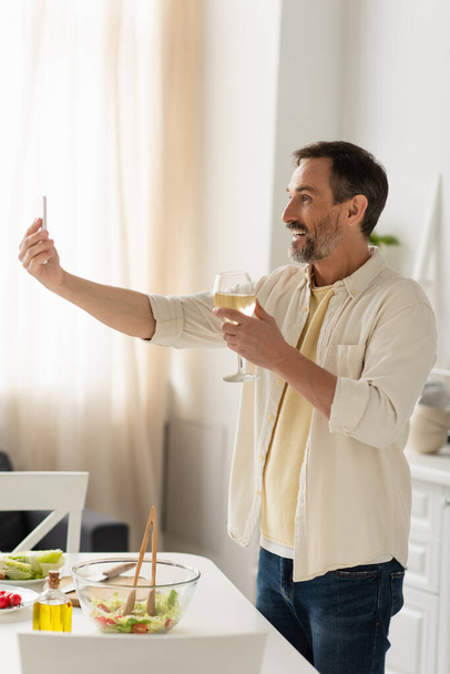 homem alegre com copo de vinho branco tomando selfie perto de legumes frescos e salada na cozinha - Foto, Imagem