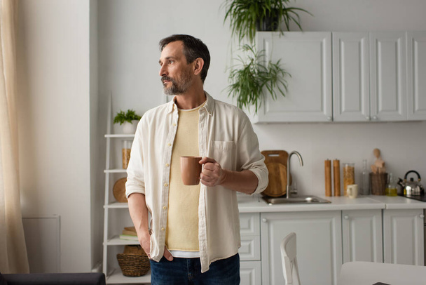 bearded man in white shirt standing with cup of tea and hand in pocket while looking away in kitchen - Foto, afbeelding