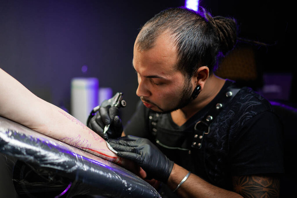 Portrait of man tattoo master with dreadlocks showing process of creation tattoo on female body under lamp light. Professional artist working in salon. High quality photo - Fotoğraf, Görsel
