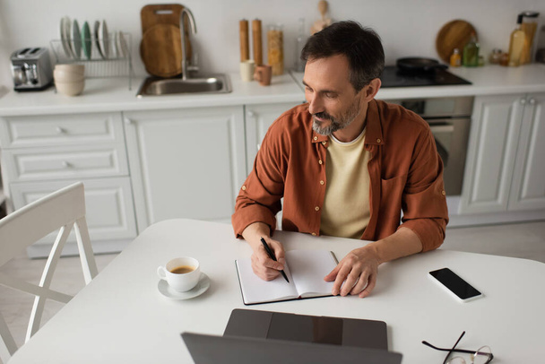 bearded man writing in notebook near laptop and coffee cup while working in kitchen - Photo, image
