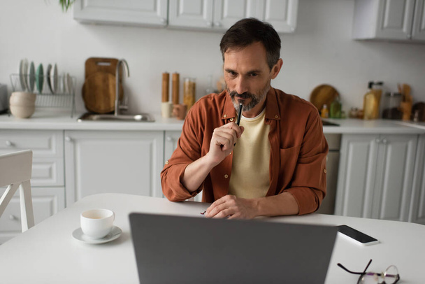 hombre pensativo con pluma mirando portátil cerca de taza de café y teléfono inteligente con pantalla en blanco - Foto, Imagen