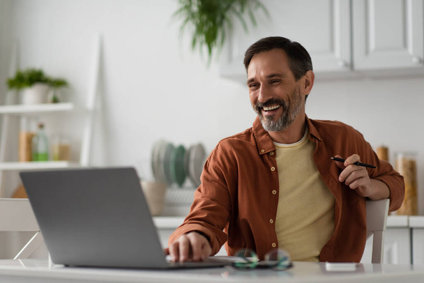 happy man working in kitchen and laughing while looking at blurred laptop - Photo, Image