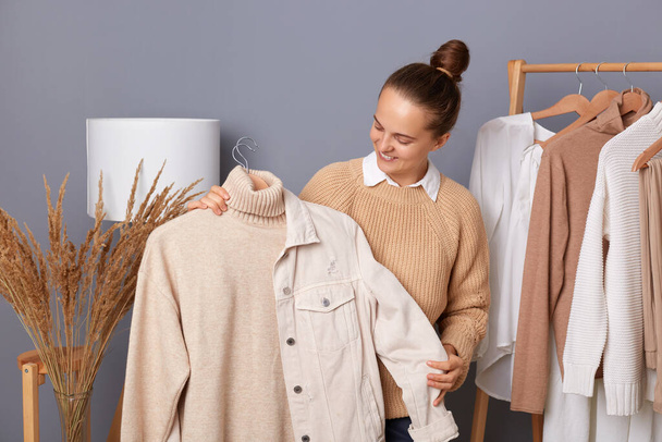 Lovely cheerful woman, fashion stylist standing near clothes hang on shelf, holding designer shirt and jacket, smiling while posing in wardrobe, combines clothes to make trendy look. - Foto, immagini