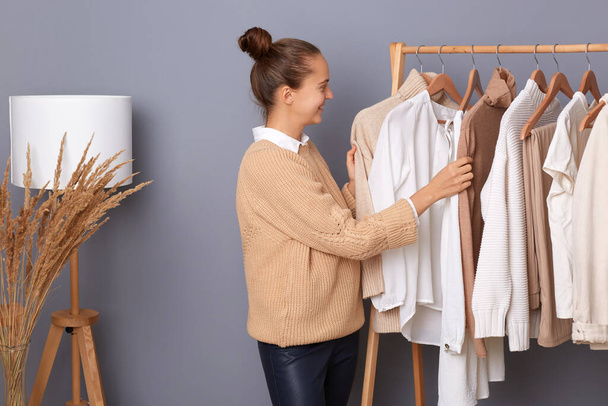 Young adult happy woman with bun hairstyle professional expert fashion stylist standing near clothes hang on shelf in designer store, selling trendy outfit in shop boutique. - Fotoğraf, Görsel