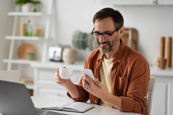 cheerful man with coffee cup looking at smartphone near empty notebook and blurred laptop - Foto, immagini