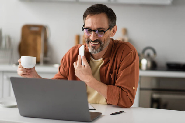 улыбающийся мужчина в очках, держащий в руках мобильный телефон и кофейную чашку, глядя на размытый ноутбук  - Фото, изображение