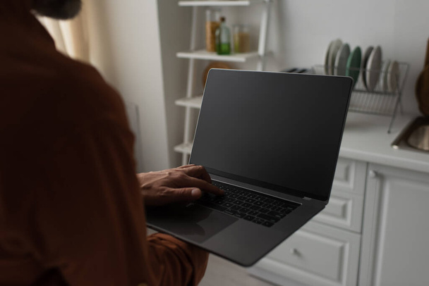 gedeeltelijke weergave van wazig man houden laptop met blanco scherm in de keuken - Foto, afbeelding