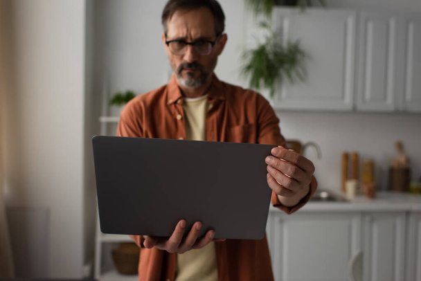 worried man in eyeglasses looking at laptop in blurred kitchen - 写真・画像