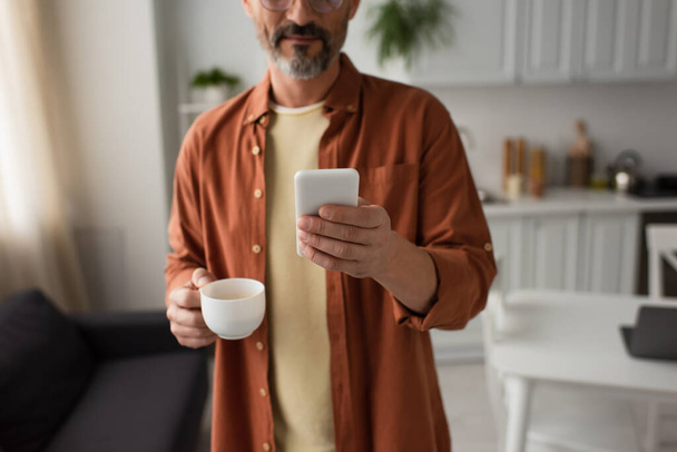 καλλιεργημένη άποψη του γενειοφόρου άνδρα σε καφέ πουκάμισο κρατώντας smartphone και φλιτζάνι καφέ στην κουζίνα - Φωτογραφία, εικόνα