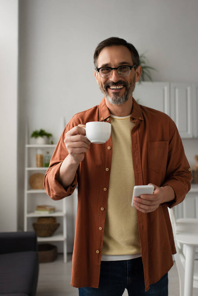 boldog férfi szemüveges kezében kávéscsésze és mobiltelefon, miközben nézi a kamera a konyhában - Fotó, kép