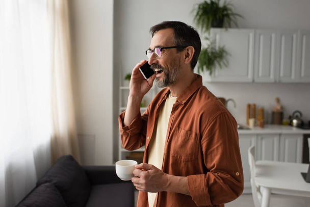 man in brown shirt and eyeglasses holding coffee cup and talking on smartphone in kitchen - Foto, afbeelding