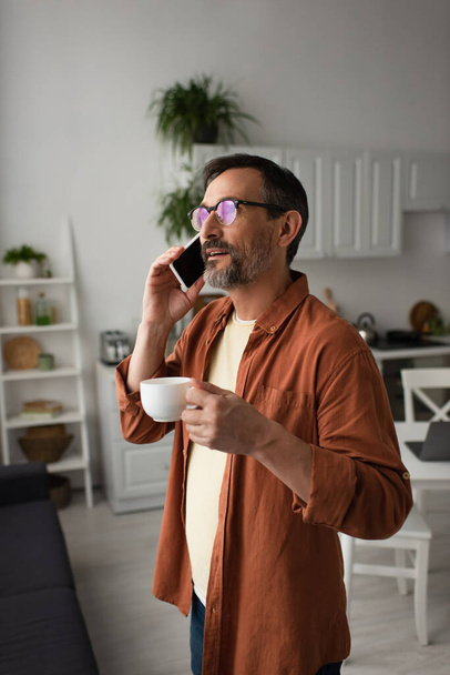 bearded man in eyeglasses talking on smartphone and holding coffee cup in kitchen - Фото, зображення