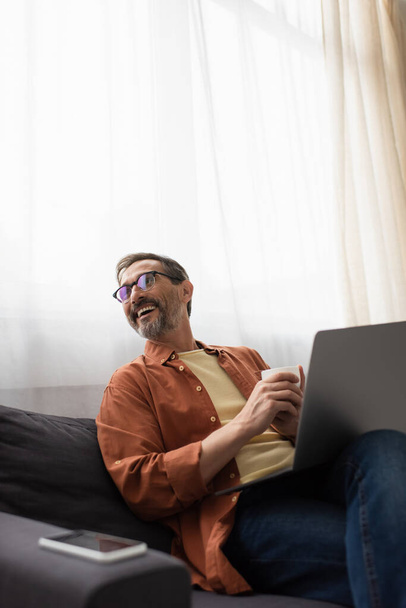 joyful man in eyeglasses looking away while sitting on couch with laptop and coffee cup - Foto, afbeelding