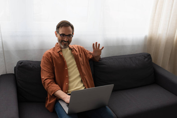 joyful bearded man waving hand during video call on laptop while sitting on sofa - Photo, Image