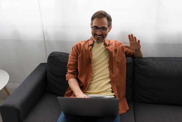 boldog szakállas férfi szemüvegben ül a kanapén, és integet kezét videohívás közben a laptop - Fotó, kép