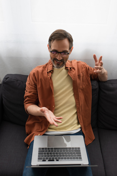 high angle view of smiling man in eyeglasses pointing at laptop during video chat on couch at home - Фото, изображение
