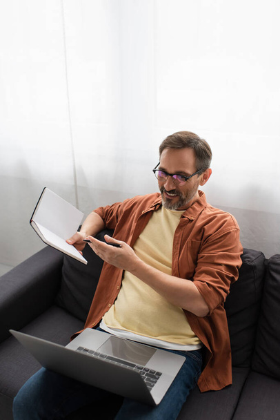 smiling man in eyeglasses pointing at empty notebook during video call on laptop at home - Photo, image