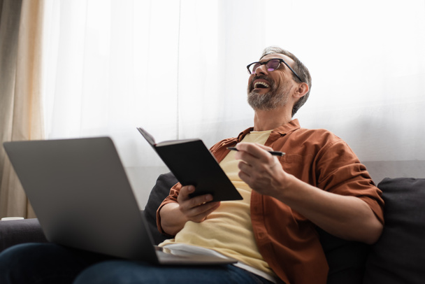 excited man in eyeglasses holding pen and notebook while laughing on couch near laptop - Fotoğraf, Görsel