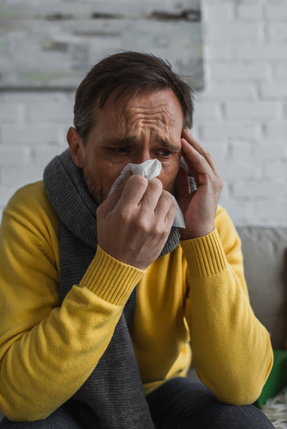 больной человек в теплом шарфе чихает в бумажную салфетку и страдает от мигрени дома - Фото, изображение