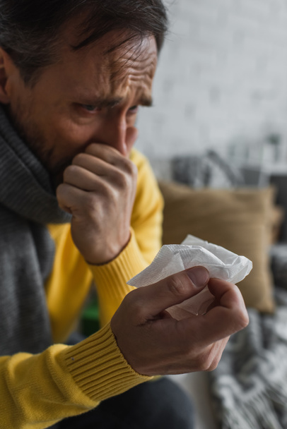 άρρωστος άνθρωπος με ζεστό κασκόλ κρατώντας χαρτοπετσέτα και φτάρνισμα στο σπίτι - Φωτογραφία, εικόνα
