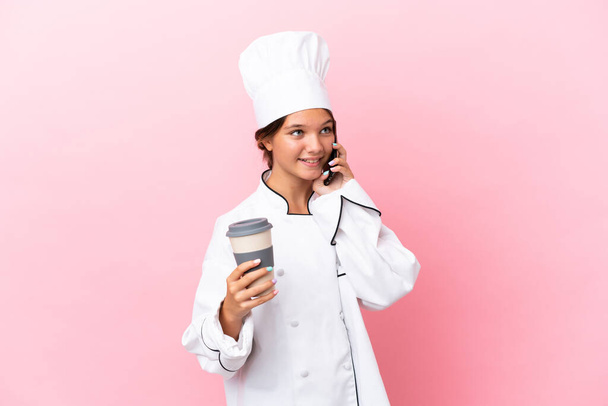 Piccola cuoca caucasica isolata su sfondo rosa con caffè da portare via e un cellulare - Foto, immagini