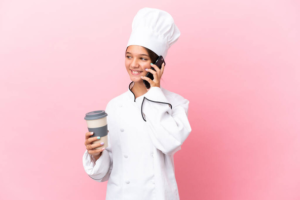 Pequena menina chef caucasiano isolado no fundo rosa segurando café para levar e um celular - Foto, Imagem