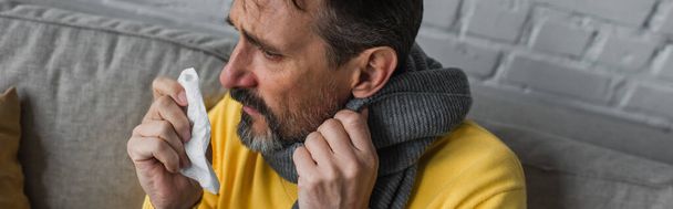 vista de ángulo alto del hombre con dolor de garganta y secreción nasal sosteniendo servilleta de papel mientras está sentado en una bufanda caliente, pancarta - Foto, Imagen