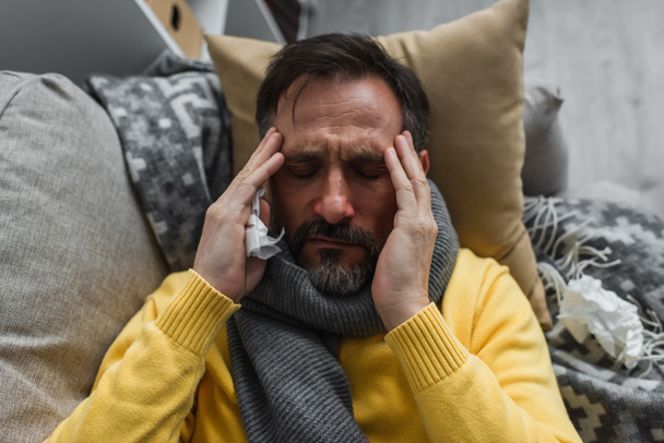 top view of sick man in warm scarf touching head while lying on couch and suffering from migraine - Photo, Image