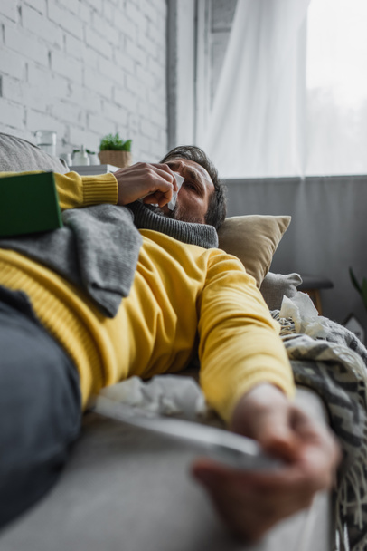 Hastalıklı bir adam ön planda bulanık bir şekilde kanepede yatarken kağıt peçeteye hapşırıyor. - Fotoğraf, Görsel