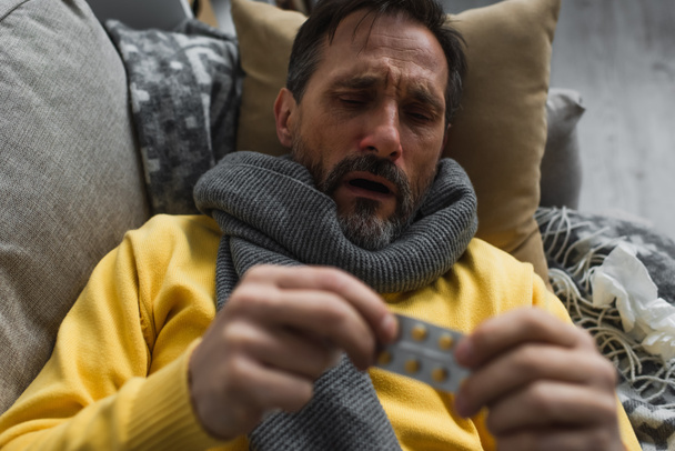 άρρωστος άνθρωπος με ζεστό κασκόλ κρατώντας θολή χάπια, ενώ βρίσκεται στον καναπέ - Φωτογραφία, εικόνα