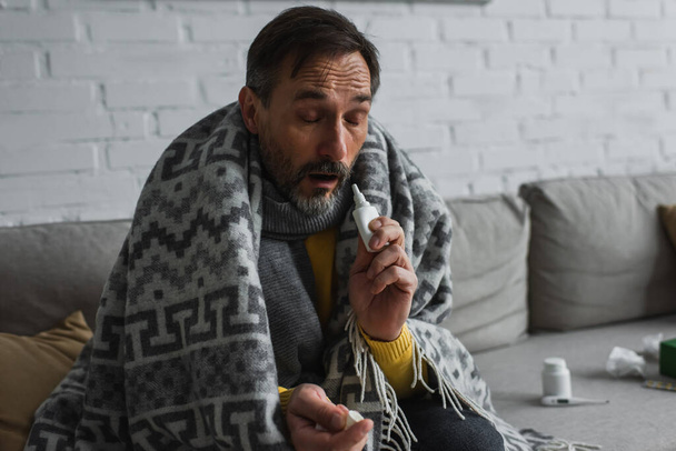 ill man with closed eyes sitting under warm blanket and holding nasal spray - Fotoğraf, Görsel