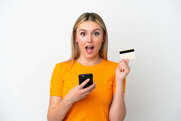 Jeune femme caucasienne isolée sur fond blanc achetant avec le mobile et tenant une carte de crédit avec expression surprise - Photo, image