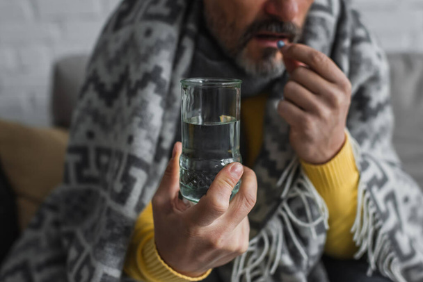 cropped view of sick man under warm blanket holding glass of water and taking pill on blurred background - Foto, imagen